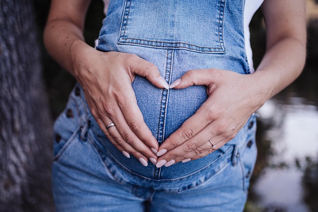 pregnant woman with fingers in the shape of a heart wearing blue jean overalls