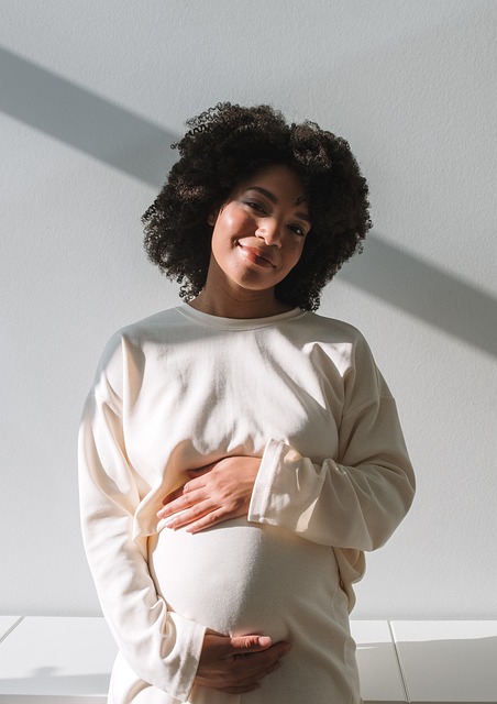 pregnant african american woman wearing white sweater standing on white tile in front of a white wall with hair in the style of an afro no makeup and natural nails